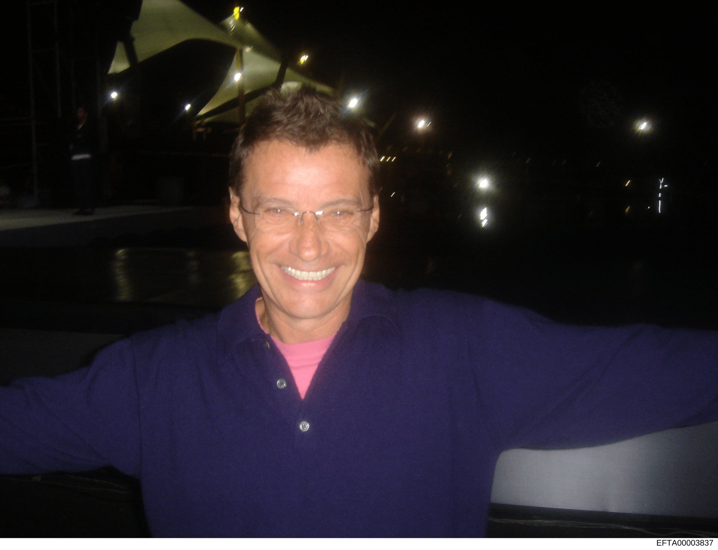 This is a photograph of a man wearing glasses and a dark blue jacket with a pink shirt underneath, photographed at night in front of what appears to be a private aircraft or hangar with illuminated fixtures visible in the background. The image is inc