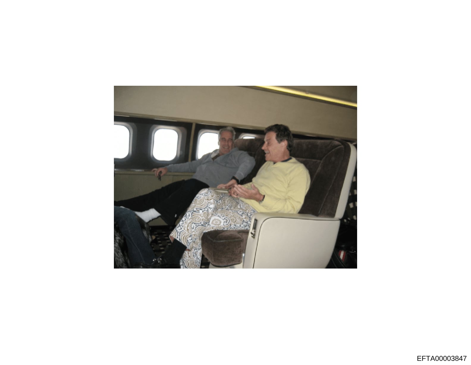 This photograph shows two men seated in the luxury interior of a private aircraft. The image appears to be documentary evidence capturing individuals aboard a private jet, with upholstered seating and cabin windows visible. The photo is potentially r