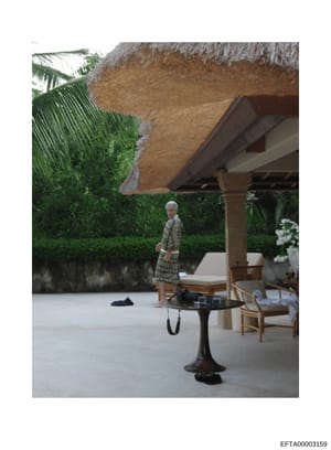 This photograph shows an outdoor patio or terrace area at what appears to be a luxury tropical property. The image features a person standing under a large thatched roof structure, with upscale outdoor furnishings including lounge chairs, a pedestal 