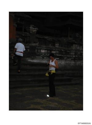 This photograph depicts two people in an outdoor setting at night or low-light conditions. One person wears a white shirt and red belt, while another wears a white sleeveless top and yellow shorts. The setting appears to be near a dark structure or b