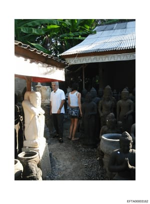 This photograph shows two adult males and a female visitor in what appears to be an art gallery, workshop, or sculpture studio setting. The space contains multiple stone sculptures and statues, with a metal roof structure. The image documents what ap