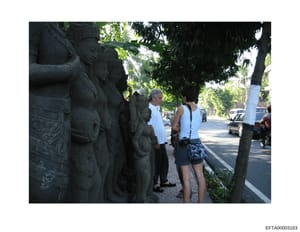 This photograph shows a street scene featuring decorative stone statuary on the left side of the frame, with pedestrians visible on a sidewalk. The image appears to be taken in a tropical or subtropical location with lush tree coverage overhead. The 