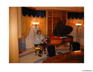 This photograph shows the interior of an elegant, formally decorated room featuring a grand piano. An older woman in gray clothing is seated at the piano. The room displays high-end furnishings including ornate curtains in gold and navy, decorative w
