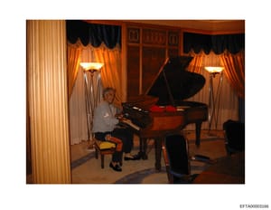 This photograph shows an elegantly decorated formal music room or salon featuring two grand pianos, ornate window treatments with gold and black drapes, matching brass floor lamps, and classical interior design. A person is seated at one of the piano