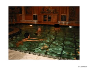 This photograph shows an indoor swimming pool with luxurious wood-paneled walls and decorative artwork. Multiple individuals are visible in the illuminated turquoise water. The image appears to be documentation of a property's interior amenities and 