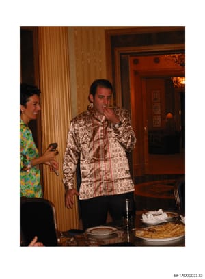 This photograph shows two people at what appears to be a formal indoor dining setting. The image depicts an interior space with classical architectural elements, including fluted columns and decorative wooden paneling. A man in a patterned silk jacke