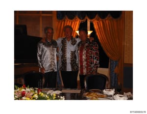 A color photograph showing three adult men wearing distinctive patterned and ornate jackets at what appears to be a formal indoor event or dinner. The setting features elegant furnishings including orange velvet curtains with decorative trim, wooden 