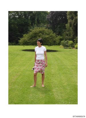 A color photograph showing a woman standing on a large manicured lawn with ornamental landscaping and trees in the background. The image appears to be taken at a residential estate property. The photograph's inclusion in the investigation files sugge