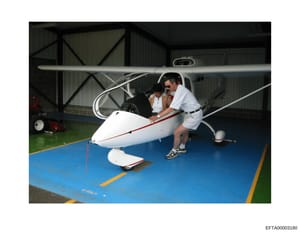 This photograph shows two people near a small white, blue, and red aircraft inside a metal-frame hangar. The aircraft appears to be a personal or recreational glider or small plane. The image is dated by its Bates number EFTA00003180 and may be relev