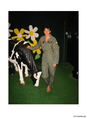 This is a personal photograph showing a woman wearing a U.S. Air Force flight suit standing next to a black and white decorative cow figure against a green background with oversized colorful flowers. The image appears to be a candid or recreational p