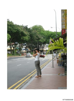 This is a color photograph showing a woman standing on a sidewalk in what appears to be a tropical or subtropical urban location, likely in Asia based on visible signage and architecture. The image shows a street scene with parked cars, palm trees, a