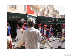 This photograph shows a daytime street scene at what appears to be a public festival or parade in a European city, likely France based on storefront signage. Multiple people wearing red and blue clothing are visible, suggesting organized participants