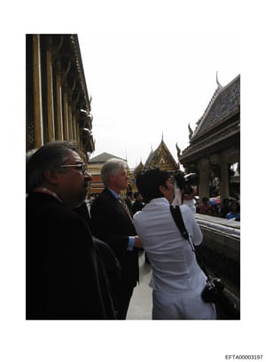 This photograph shows a group of individuals visiting a Thai Buddhist temple, identifiable by the distinctive golden spires and ornate architectural details characteristic of Thai temple design. The image appears to be documentary in nature, capturin