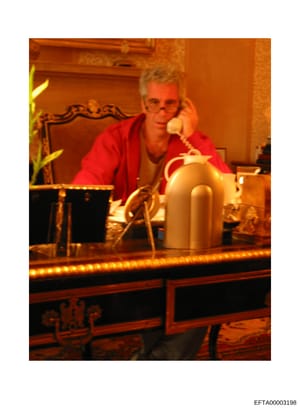This is a photograph showing a man wearing a red shirt seated at an ornate desk with decorative elements, speaking on a telephone. The image appears to be taken in an interior space with warm lighting and luxurious furnishings. The photograph is part