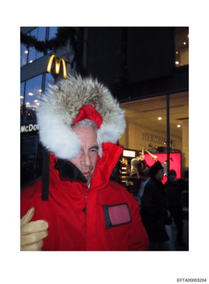 This is a photograph of a man wearing a distinctive red winter jacket with a large white fur hood and red fur trim, taken on a city street at night. The image appears to be from a public event or gathering, with McDonald's and other storefront signag