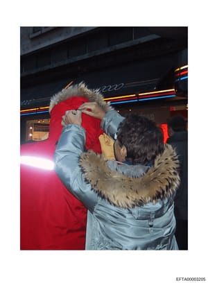 This is a photograph showing a person wearing a light blue puffy jacket with fur-lined hood and red cap/hat at what appears to be a public venue or entertainment location. The background shows neon signage and storefront elements. The photo's context