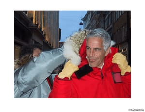 This is a photograph showing a man wearing a red winter coat with fur-lined hood and yellow gloves on an urban street with period architecture. The man appears to be posing for the camera on what looks like a European city street. The image is includ