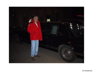 This is an evidence photograph from the Epstein investigation files showing a man wearing a distinctive red jacket with fur collar and light blue jeans standing next to a black vehicle at night. The photo appears to be taken in a parking garage or si