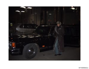 This is an evidence photograph from the Epstein investigation showing a woman standing next to a black luxury sedan on a dark urban street at night. The image appears to be surveillance or documentary photography, with the subject wearing a dark coat