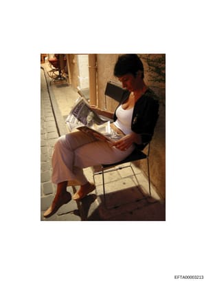 This is a casual photograph showing a woman seated in a chair in what appears to be a residential hallway or corridor, reading a newspaper. The image is undated and lacks contextual information about its relevance to the investigation. It appears to 