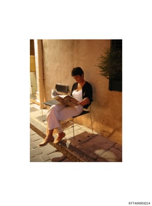 This is a color photograph showing a woman seated in a courtyard or alleyway area reading a magazine. She is dressed casually in a white top and pink/lavender pants with a black cardigan. The setting appears to be a Mediterranean or European-style re