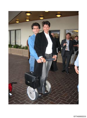 This is a photograph taken at an indoor event or venue showing two people posing together on a Segway personal transportation device. The setting appears to be a professional or corporate environment with modern lighting, planted vegetation, and othe