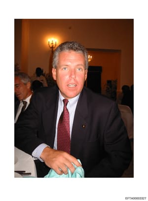 This is a color photograph of a middle-aged man wearing a dark business suit and red patterned tie at what appears to be an indoor formal event or gathering. The setting features warm lighting, a wall sconce, and other attendees visible in the backgr