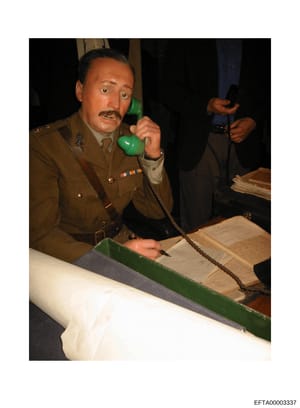 This is a photograph of a wax museum display or historical diorama depicting a military officer in a tan/khaki uniform with medals and insignia, speaking on a vintage green rotary telephone. The figure is positioned at a desk with documents and paper