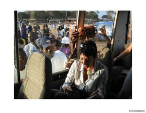 This photograph shows the interior of a transport vehicle, likely a bus or van, filled with multiple passengers. The image appears to have been taken at a community or school sports event, with athletic fields, spectators, and what appears to be a po