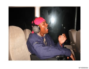 This is an evidence photograph showing a man seated in what appears to be an aircraft cabin, wearing a pink/red headband, Sony headphones, and a dark blue jacket. The photo appears to be from the Epstein investigation files and was likely taken durin