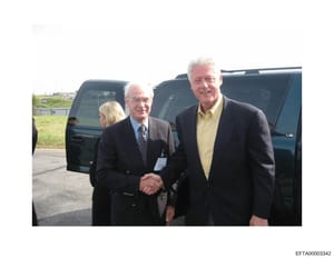 This photograph shows three men in business attire shaking hands outdoors near parked dark-colored vehicles. The man on the left wears a dark suit with a name badge/credential, the man in the center wears a dark suit with glasses, and the man on the 