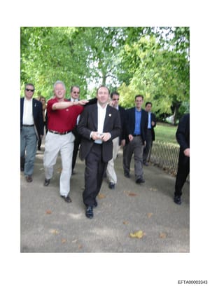 This is a color photograph showing a group of approximately 8-10 men walking together along a tree-lined path or park grounds. The image appears to be candid documentation of individuals in business and casual attire. Without additional context or id