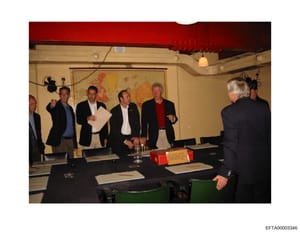 This photograph shows a formal meeting or conference taking place in an interior room with a distinctive red ceiling and cream-colored walls. Multiple men in business attire are present, appearing to be engaged in discussion around a table with paper