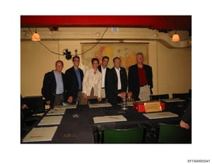 This photograph shows six men standing behind a conference table in what appears to be an official or business meeting setting. The image is undated but appears to be from the 1990s based on clothing and photo quality. One name placard is visible on 