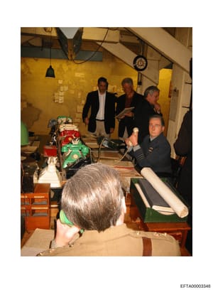 This photograph shows a group of approximately 6-7 people in what appears to be an industrial or workshop space with yellow walls. Several men in business attire are observing equipment or machinery (including red and green colored devices), while on