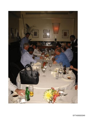 This photograph shows a formal dinner gathering of approximately 10-12 adults seated around a white tablecloth-covered table in an upscale dining establishment. The image captures a private event with professional ambiance, featuring formal place set
