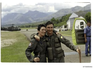 This photograph shows two military personnel in flight suits posing together at what appears to be a remote mountain airfield or landing zone. A military helicopter is visible in the background on the left, along with support buildings and a lime-gre