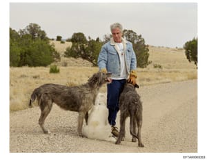 This is a color photograph showing a man with gray/white hair wearing a denim jacket and work gloves standing on a dirt road with two large dogs. The setting appears to be a rural, semi-arid area with scattered vegetation and open landscape. The phot