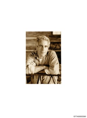 This is a sepia-toned portrait photograph showing a middle-aged man with gray/blonde hair wearing a tan/beige button-up shirt, posed with arms crossed leaning against what appears to be a railing or bar. The photograph appears to be a professional or