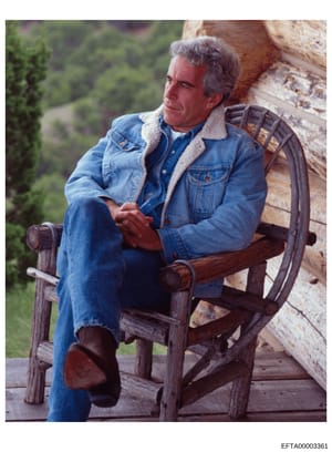 This is a color photograph showing a middle-aged man with gray hair wearing a blue denim jacket with sherpa lining, seated in a rustic wooden chair against a stone building exterior. The setting appears to be outdoors with landscaped grounds visible 