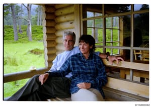 This is a color photograph depicting two individuals posing together on what appears to be a deck or porch of a rustic log cabin structure. The setting shows a forested landscape with green grass and tall trees visible in the background through large