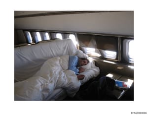 This photograph shows the interior of what appears to be a private aircraft, specifically a sleeping or bedroom area with a bed, white bedding, and windows along the fuselage. A person is visible resting on the bed. The image documents the luxury acc