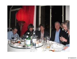 This photograph depicts five individuals seated at a round dining table in an upscale interior setting with Asian-inspired décor. The scene shows an elegant dinner gathering with place settings, wine glasses, bottles (including what appears to be Per