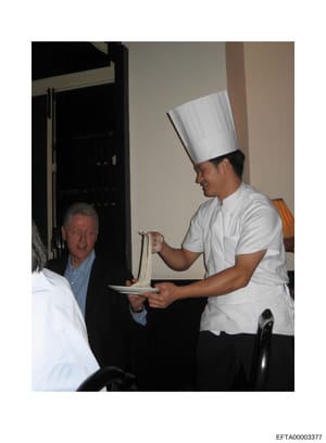 This photograph shows a chef in white uniform and tall chef's hat preparing or serving noodles to a man in a blue shirt at what appears to be an indoor culinary event or demonstration. The image appears to be evidence documentation from the investiga
