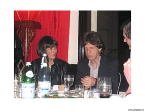 A photograph showing a formal dining scene with three visible individuals seated at a table with place settings, glasses, and bottles of Pellegrino water. The setting features elegant decor including red fabric with decorative Asian-inspired elements