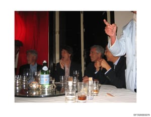 This is a photograph of what appears to be an indoor meeting or social event, showing four men seated at a table with glasses, bottles, and place settings. The setting suggests a formal or business gathering, with red curtains visible in the backgrou