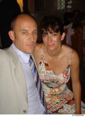 This is a photograph showing two people posing together at what appears to be an indoor social event or gathering. The man is wearing a beige blazer with a light blue shirt and striped tie, while the woman is wearing a colorful patterned sundress. Ot