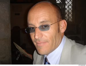 This is a portrait photograph of a middle-aged man wearing glasses, a light-colored blazer, and a blue tie with a red stripe. The photo appears to be taken indoors in a residential or institutional setting. Without additional context or identifying i