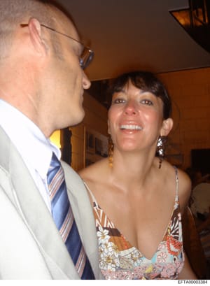This is a candid photograph showing two people at what appears to be an indoor social gathering or event. The image shows a man wearing glasses and a light-colored suit with a striped tie, and a woman with dark hair wearing a patterned dress with flo