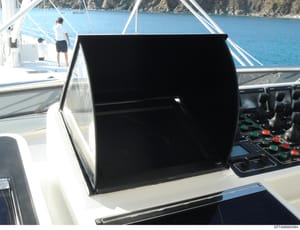 This photograph shows the interior cockpit/bridge of a luxury yacht at sea, featuring navigation and engine control equipment on the right side of the frame. The image captures two crew members on the deck area in the background, with clear blue wate